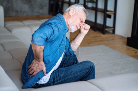 Unhappy mature man feeling bad, suffering from backache, hold his back and can't stand up from sofa. Middle aged male with discomfort and kidney disease sitting alone on couch at home