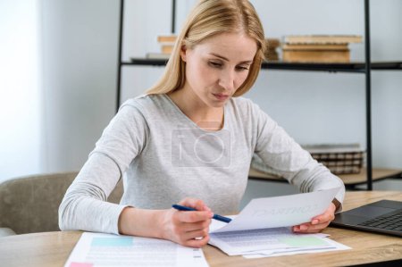 Focused woman sitting in home office, checking documents, looking looking at paper contract. Business consultant or manager reading agreement information at workplace.