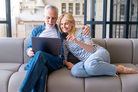 Feliz pareja de la familia madura con el ordenador portátil y la tarjeta de crédito de compras en línea desde casa. Mujer sonriente y hombre sentado en la sala de estar, abrazar en el sofá y hacer compras por Internet. Concepto de comercio electrónico.