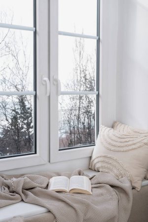 Téléchargez les photos : Plan vertical de grandes fenêtres en plastique avec vue d'hiver et rebord de fenêtre dans le salon à la maison. Endroit confortable pour s'asseoir et lire livre avec plaid confort doux et coussins. - en image libre de droit