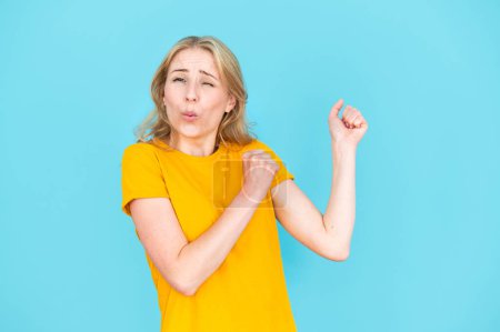Optimistic and excited young woman in yellow t-shirt dancing isolated on blue copy space background. Concept of positive emotion. Happy overjoyed female raised fists up, moving on celebration party