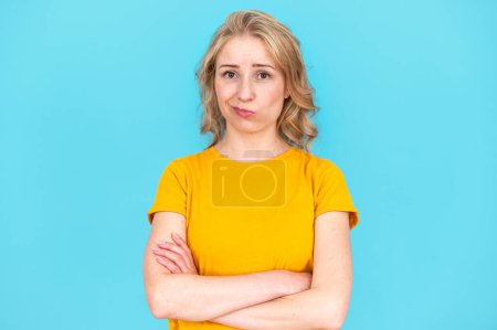 Studioaufnahme einer Frau mit verschränkten Armen, die in die Kamera blickt, mit zweifelndem und unentschlossenem Gesichtsausdruck. Verwirrte junge Frau posiert isoliert auf blauem Hintergrund.