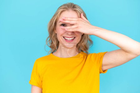 Photo for Smiling woman covering face with hand and peeping out fingers isolated on blue background. Looking at camera. Spying on something interesting. Special offer. Copy space. - Royalty Free Image