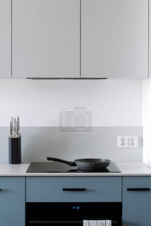 Vertical shot of modern elegant kitchen interior with built-in household equipment. Grey countertop with induction hob with cupboard above. Blue drawers next to electric oven.
