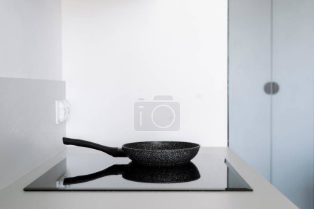 Großaufnahme von Elektroherd mit Pfanne auf sauberer schwarzer Keramikoberfläche gegen weiße Wand in der modernen Küche. Eingebaute Haushaltsgeräte für zu Hause. Kopierraum.
