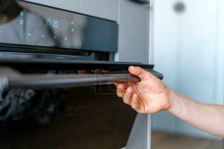 Photo for Selective focus on woman hand holding and opening glass black door of built-in electric oven in modern kitchen. Control panel with digital timer and buttons on domestic appliance for baking. - Royalty Free Image