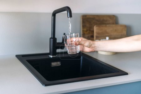 Selective focus on female hand holding glass and pouring fresh drink water from kitchen stainless black steel faucet above ceramic sink on white counter top. Copy space