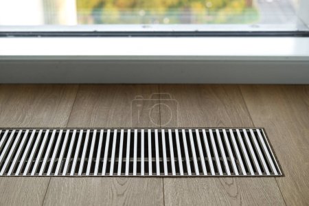 Close up of wooden floor with chrome ventilation grid of central underfloor heating system against window in apartment. Energy consumption in cold season. Concept of smart home.