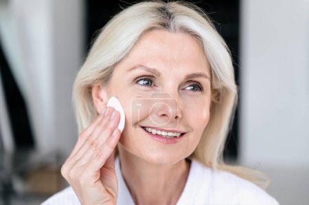 Photo for Mature woman with healthy smooth skin applying hyaluronic moisturizing toner with cotton pads at home bathroom. Daily hygiene procedures and skincare routine. - Royalty Free Image