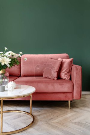 Photo for Vertical shot of cozy living room interior with red comfortable sofa with cushions and coffee table with marble countertop and vase with flowers. Wooden floor and green wall in apartment. - Royalty Free Image