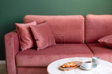 Photo for Selective focus on bamboo tray with bakery, cup with morning tea on coffee table with marble top next to coral couch against green wall in living room in classic style at apartment. Breakfast concept - Royalty Free Image