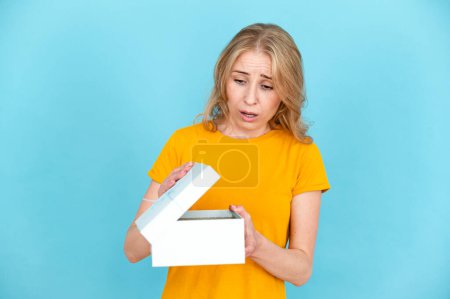 Upset shocked woman in yellow t-shirt opening gift box isolated on blue background. Disappointed sad female unpacking present.