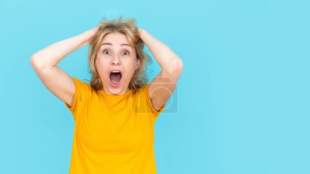 Woman holding head with hand and feeling panic and shock standing on blue studio wall background. Frustrated student or freelancer showing negative emotion, worrying, banner view, copy space