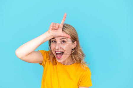 Young woman in yellow t-shirt showing loser gesture by fingers on forehead, teasing and mocking isolated on blue background. Female making fun of people and laughing