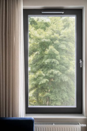 Foto de Imagen vertical de una ventana de pvc cerrada en negro con una increíble vista al bosque. Acogedor salón interior con cortinas beige. Verano, concepto de primavera. - Imagen libre de derechos