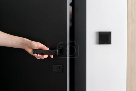 Photo for Cropped shot of woman hand open black door to the living space. Female use mechanical metal handle to access in hotel room or apartment - Royalty Free Image
