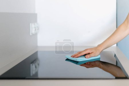 Frau mit blauem Mikrofasertuch reibt Glaskeramikherd in der Küche. Hausfrau poliert ihren modernen und neuen Elektroherd. Selektiver Fokus, Kopierraum Hintergrund. Haushaltsgerätekonzepte
