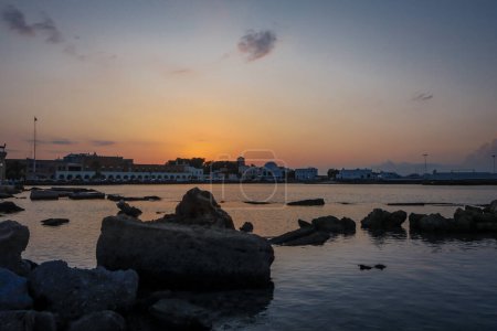 Téléchargez les photos : Rhodes, Grèce - 27 mai 2023 : Coucher de soleil à Mandraki Rhodes Marina. Palais des gouverneurs bâtiment en arrière-plan, pierres de plage au premier plan. - en image libre de droit