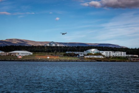 Téléchargez les photos : Reykjavik, Islande - 25 septembre 2023 : Paysage avec l'aéroport domestique de Reykjavik et le bâtiment Perlan. Un petit avion dans l'air, baie d'eau au premier plan. - en image libre de droit
