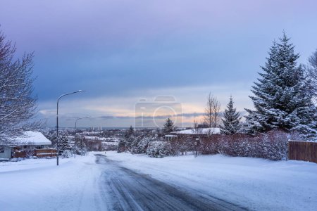 Foto de Reikiavik, Islandia - 15 de enero de 2024: Calle nevada vacía en los suburbios de Reikiavik en un oscuro día de invierno. Edificios residenciales, coches conduciendo por la carretera. Gris, nubes tormentosas en el cielo. - Imagen libre de derechos