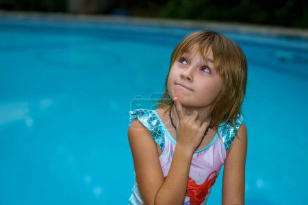 girl in a swimsuit 8-9 years old near the pool with a doubtful and thoughtful expression on her face.