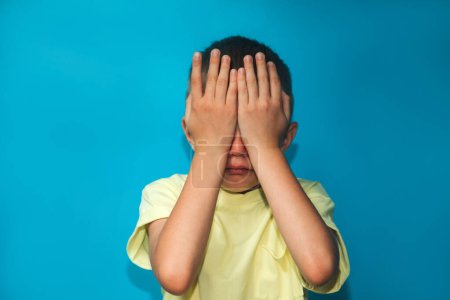 Foto de The boy is upset and crying with close eyes by hands on a blue background. Portrait of a child schoolboy. Front view - Imagen libre de derechos