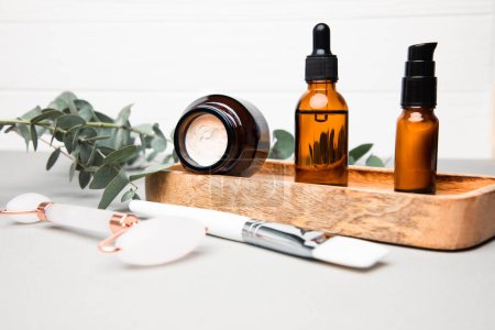 Amber bottles with facial cosmetics, face roller and brush for mask on a tray with a branch of eucalyptus on the gray concrete background. Front view