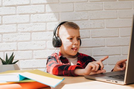 Photo for A school-age boy in black headphones sits at a table with a laptop. Children and gadgets. Front view - Royalty Free Image