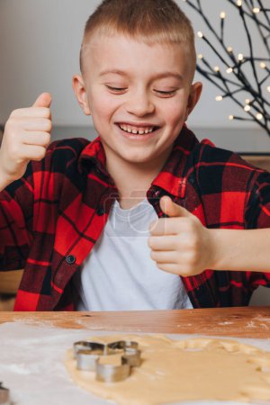 Foto de Un niño con una camisa a cuadros roja en una mesa de madera en la cocina va a hacer galletas sosteniendo un cortador de galletas. Vista frontal - Imagen libre de derechos