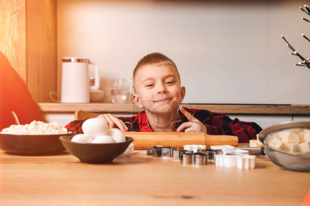 Foto de Un niño con una camisa a cuadros roja en una mesa de madera en la cocina va a hacer galletas sosteniendo un cortador de galletas. Vista frontal - Imagen libre de derechos