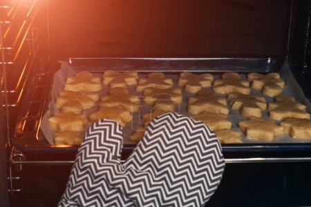 Foto de La mano de cerca en una manopla gris pone en el horno una bandeja para hornear con galletas de shortbread de diferentes formas. Preparándose para la Navidad. Cocinar galletas en casa. Vista frontal - Imagen libre de derechos