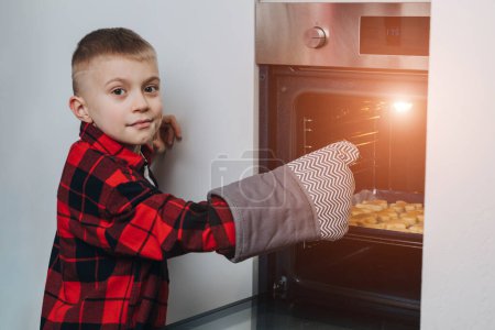 Foto de Un niño con una camisa roja pone una bandeja para hornear con galletas de Navidad en el horno. Preparándose para la Navidad. Cocinar galletas en casa. Vista frontal - Imagen libre de derechos