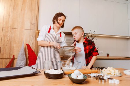 Foto de Mamá e hijo están amasando masa para galletas en una mesa de madera en la cocina. Cocinar postres en casa. Actividades conjuntas con niños. Vista frontal - Imagen libre de derechos