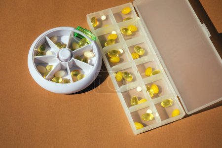 Kapseln mit Vitaminen, Vitamin D und Omega im Medikamentenbehälter auf braunem Hintergrund. Ansicht von oben