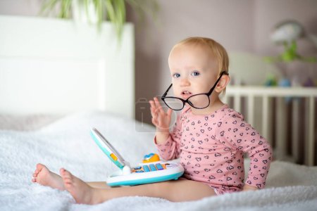 Cute toddler in glasses sitting on bed with a toy laptop