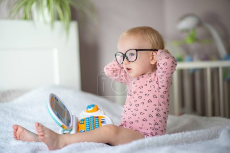 Cute toddler in glasses sitting on bed with a toy laptop