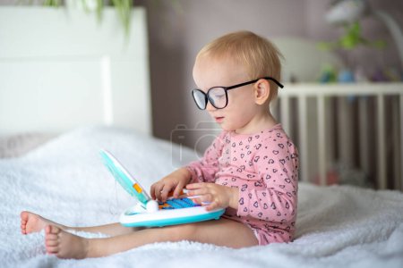 Curious toddler with glasses looking at a toy laptop on a bed