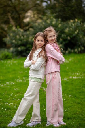 Zwei junge Mädchen in pastellfarbenen Outfits stehen Rücken an Rücken vor grünem Gartenhintergrund