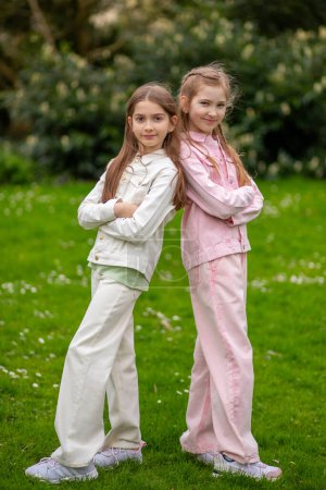 Zwei junge Mädchen in pastellfarbenen Outfits stehen Rücken an Rücken vor grünem Gartenhintergrund