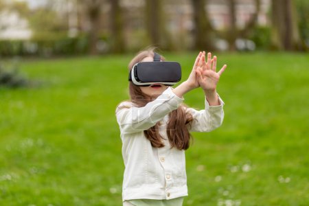 Girl in virtual reality headset raises hands in a park.