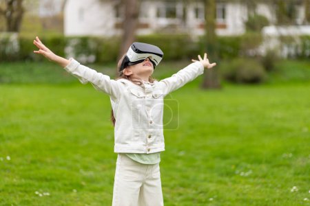 Girl in virtual reality headset raises hands in a park.