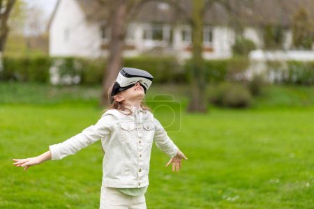 Girl in virtual reality headset with arms outstretched in a park.