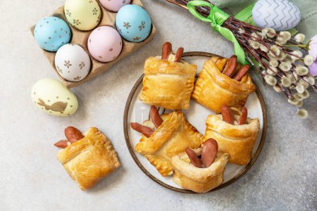 Buns in the form of an Easter rabbit from sausage and cheese in a yeast dough with colored eggs on a gray stone background. Traditional Easter symbols and food. View from above. 