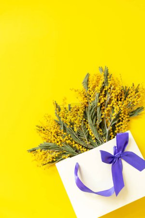 Téléchargez les photos : Mimosa fleurit sac en papier cadeau sur fond jaune. Concept de printemps, composition des fleurs. Carte de vœux. Vue d'en haut. Espace de copie. - en image libre de droit