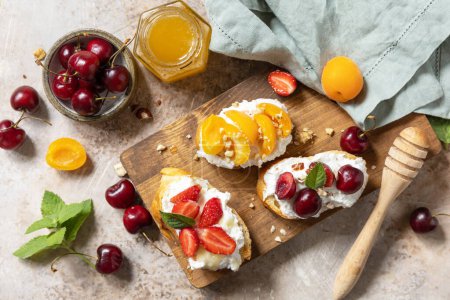 Foto de Bayas tostadas desayuno, comida saludable. Sandwich con cereza, fresas, queso suave y miel sobre tabla de madera sobre un fondo de piedra. Vista desde arriba. - Imagen libre de derechos