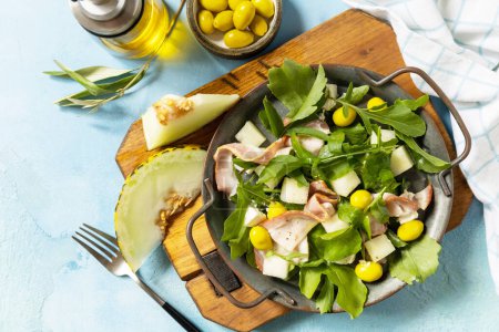Photo for Summer mediterranean salad with melon, bacon, olives, white cheese and arugula on blue stone background. Traditional Spanish and Italian appetizer. View from above. - Royalty Free Image