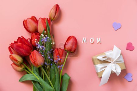 Tulipanes rojos con caja de regalo y corazones pequeños, texto escrito a mano "mamá" sobre fondo rosa pastel con espacio vacío. Concepto de celebración del Día de la Madre. Vista desde arriba.