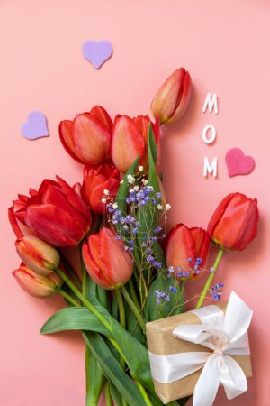 Tulipanes rojos con caja de regalo y corazones pequeños, texto escrito a mano "mamá" sobre fondo rosa pastel con espacio vacío. Concepto de celebración del Día de la Madre. Vista desde arriba.