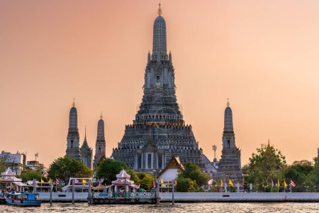 Foto de Wat Arun stupa, o el Templo del Amanecer, un hito significativo de Bangkok, Tailandia, se encuentra prominentemente a lo largo del río Chao Phraya, con un hermoso cielo anaranjado al atardecer. - Imagen libre de derechos