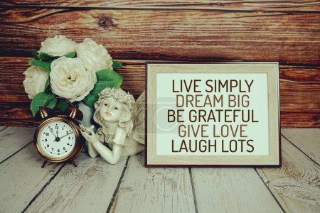 Photo for Inspirational quotes of "live simply,dream big, be grateful, give love, laugh lots" text message on wooden board with flower, alarm clock and ceramic pretty doll decoration - Royalty Free Image